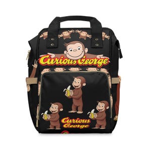 Puede incluir: Mochila negra con ilustraciones de Curious George. Tiene un asa superior, correas ajustables y múltiples compartimentos. El diseño incluye el logotipo de Curious George e imágenes del mono sosteniendo plátanos.