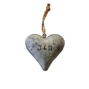 May include: A heart-shaped metal ornament with a weathered gray finish and a rustic texture. The letters J & B are stamped in black in the center. A brown jute twine loop is attached to a metal ring at the top for hanging purposes.