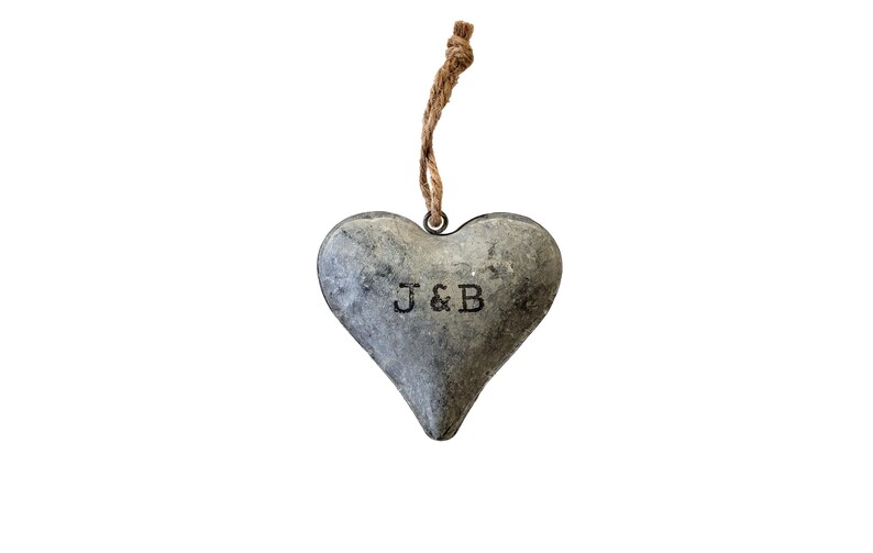 May include: A heart-shaped metal ornament with a weathered gray finish and a rustic texture. The letters J & B are stamped in black in the center. A brown jute twine loop is attached to a metal ring at the top for hanging purposes.