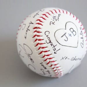 Puede incluir: B&eacute;isbol blanco con costuras rojas, cubierto de mensajes escritos a mano y un coraz&oacute;n con las iniciales "J+B".
