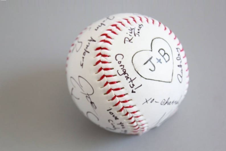 Puede incluir: B&eacute;isbol blanco con costuras rojas, cubierto de mensajes escritos a mano y un coraz&oacute;n con las iniciales "J+B".