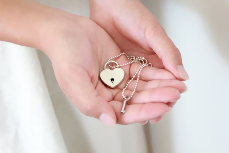 Pode incluir: Um colar com pingente em forma de cora&ccedil;&atilde;o prateado com uma chave pequena. O pingente em forma de cora&ccedil;&atilde;o tem um buraco de fechadura no centro. O colar &eacute; segurado em m&atilde;os abertas contra um fundo branco macio.