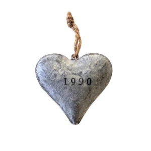May include: A heart-shaped ornament crafted from grey, textured metal with a weathered finish. The number 1990 is printed in black ink on the front center. A short, knotted piece of brown jute twine is attached to the top for hanging purposes.
