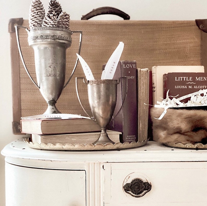 May include: Two silver trophy cups with a stack of books and a fur hat on a white dresser. The books have titles like "Love" and "Little Men".