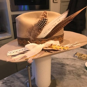 May include: A tan felt cowboy hat with a brown leather band and a decorative pattern. The hat is adorned with feathers and a white ribbon. The hat is on a stand.