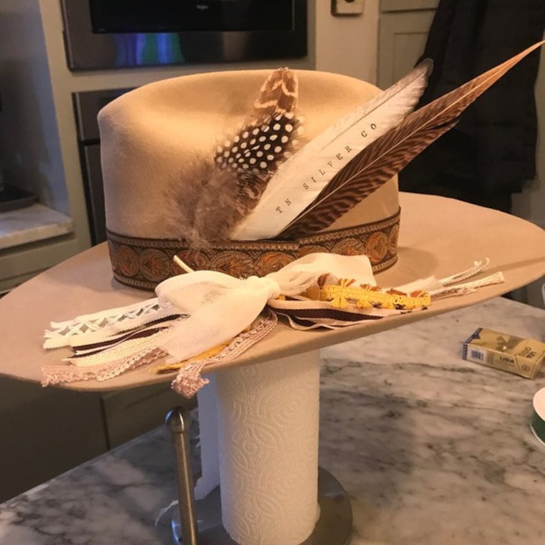 May include: A tan felt cowboy hat with a brown leather band and a decorative pattern. The hat is adorned with feathers and a white ribbon. The hat is on a stand.