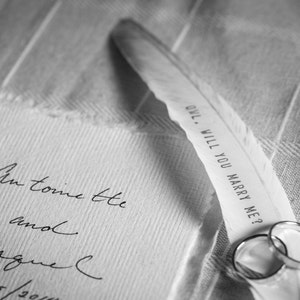 May include: A white feather with the text "Owl. Will you marry me?" written on it. A piece of paper with the text "Antoinette and Raquel 8/15/201" is visible in the background. Two silver rings are on the feather.