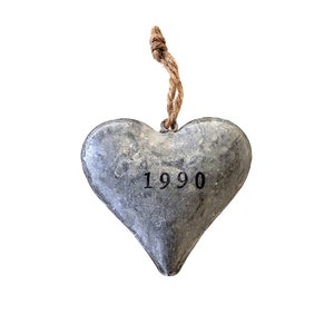 May include: A heart-shaped ornament made of gray metal with a textured, weathered finish. The number 1990 is stamped in black ink in the center of the heart. A short, knotted piece of brown jute twine is attached to the top for hanging purposes.
