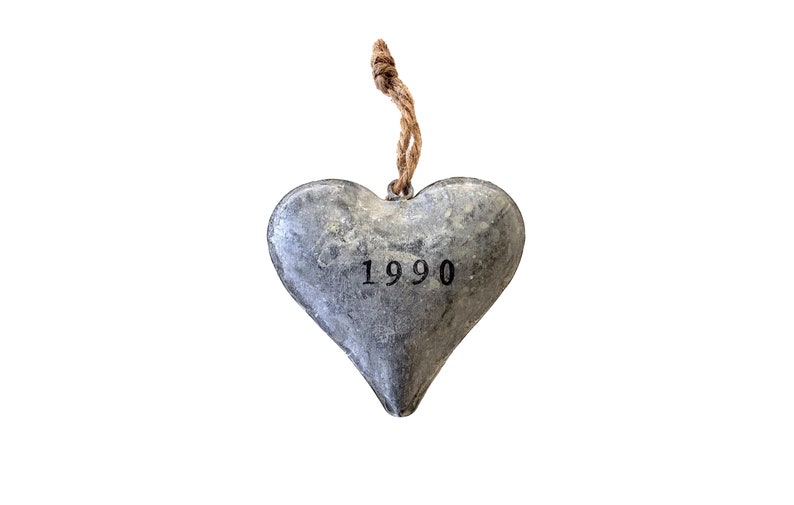May include: A heart-shaped ornament made of gray metal with a textured, weathered finish. The number 1990 is stamped in black ink in the center of the heart. A short, knotted piece of brown jute twine is attached to the top for hanging purposes.