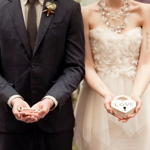 May include: A couple holding a white heart-shaped box with the word "LOVE" on it and a silver key. The woman is wearing a white strapless dress with floral embellishments and a silver necklace. The man is wearing a dark gray suit with a white shirt and a dark tie.