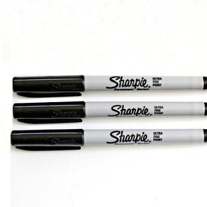 Puede incluir: Tres marcadores permanentes negros Sharpie con barriles grises. Cada marcador dice "Sharpie Ultra Fine Point Permanent Marker" en el barril.