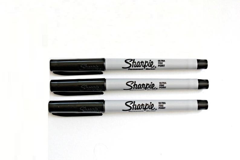 Puede incluir: Tres marcadores permanentes negros Sharpie con barriles grises. Cada marcador dice "Sharpie Ultra Fine Point Permanent Marker" en el barril.