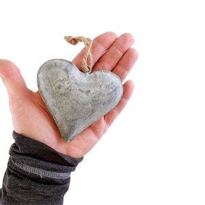 May include: A heart-shaped ornament crafted from grey metal with a weathered, textured surface. The heart features a small metal loop at the top, which is attached to a short piece of natural tan jute twine for hanging purposes.