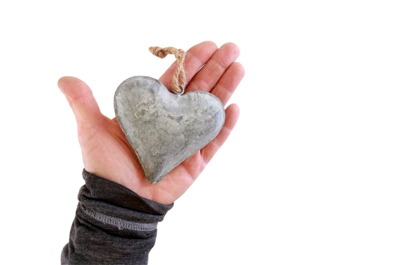 May include: A heart-shaped ornament crafted from grey metal with a weathered, textured surface. The heart features a small metal loop at the top, which is attached to a short piece of natural tan jute twine for hanging purposes.