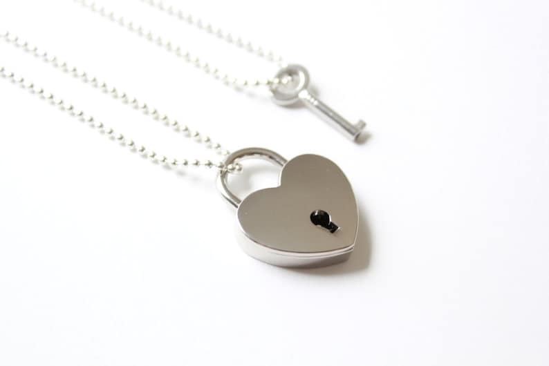 Pode incluir: Um colar com pingente de cadeado prateado em forma de cora&ccedil;&atilde;o com um colar com pingente de chave. O pingente em forma de cora&ccedil;&atilde;o tem um buraco de fechadura. Ambos os colares t&ecirc;m uma corrente de bolas. O fundo &eacute; branco.