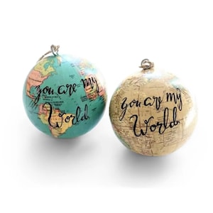 May include: Two globe ornaments, one teal and one beige, with the words "you are my world" in black script. The teal globe has a map design, while the beige globe has a vintage map design. Both have a metal loop for hanging.