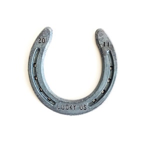 May include: A silver horseshoe with the numbers "20" and "11" stamped on each side. The horseshoe has the words "LUCKY US" stamped on the bottom.
