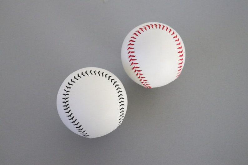 Puede incluir: Dos pelotas de b&eacute;isbol blancas con costuras rojas y negras. Una pelota de b&eacute;isbol tiene costuras rojas y la otra tiene costuras negras.