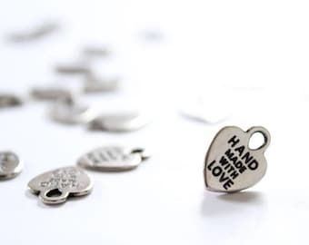 May include: Silver heart-shaped charms with the words "HAND MADE WITH LOVE" engraved on them. The charms have a small loop at the top for attaching to jewelry or other crafts. Several charms are scattered on a white surface.