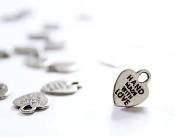 May include: A collection of small, silver-colored heart-shaped metal charms. Each charm features a loop at the top for attachment and the text "HAND MADE WITH LOVE" embossed in black lettering on the front surface of the heart-shaped metal piece.