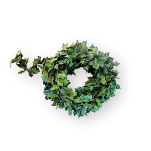 May include: A coiled garland of artificial green ivy leaves featuring a dense arrangement of pointed, multi-tonal foliage. The vine is wound into a circular shape with a single loose end extending to the left, showcasing realistic leaf textures.