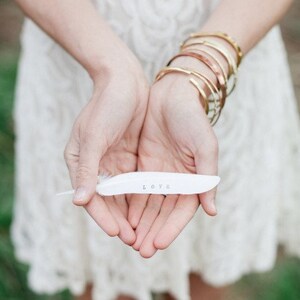 May include: A white feather with the word "LOVE" written on it, held in the hands of a person wearing a white lace dress and gold bracelets.