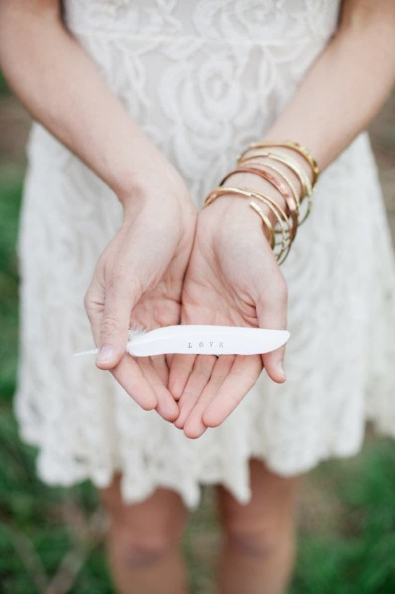 May include: A white feather with the word "LOVE" written on it, held in the hands of a person wearing a white lace dress and gold bracelets.
