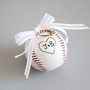 Puede incluir: B&eacute;isbol blanco con costuras rojas y un coraz&oacute;n con las iniciales "J+B" escritas en su interior. Un anillo de oro se coloca en la parte superior de la pelota, que est&aacute; atada con una cinta blanca.