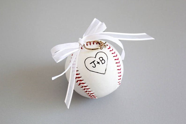 Puede incluir: B&eacute;isbol blanco con costuras rojas y un coraz&oacute;n con las iniciales "J+B" escritas en su interior. Un anillo de oro se coloca en la parte superior de la pelota, que est&aacute; atada con una cinta blanca.