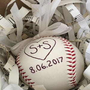 Puede incluir: Una pelota de b&eacute;isbol blanca con costuras rojas, decorada con un coraz&oacute;n y las iniciales "S+S" y la fecha "8.06.2017" escrita con un marcador negro.