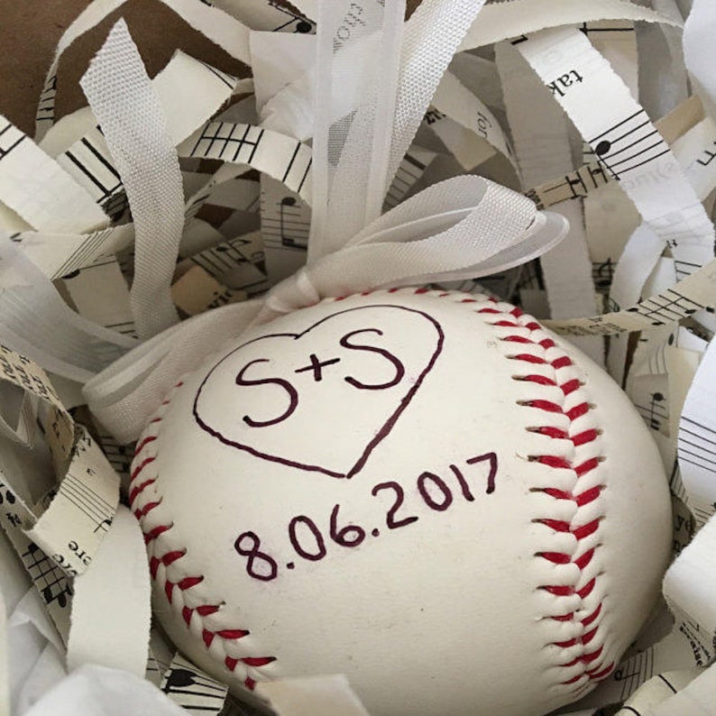 Puede incluir: Una pelota de b&eacute;isbol blanca con costuras rojas, decorada con un coraz&oacute;n y las iniciales "S+S" y la fecha "8.06.2017" escrita con un marcador negro.