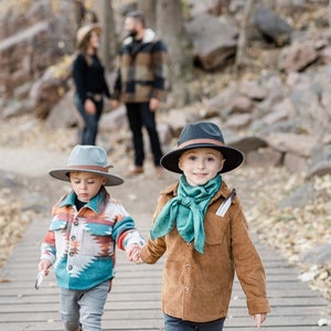 May include: Two young boys wearing hats walk along a wooden path in a forest. The boy on the left is wearing a gray hat and a blue, orange, and white patterned shirt. The boy on the right is wearing a black hat and a brown jacket with a teal scarf.
