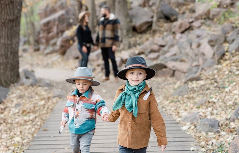 May include: Two young boys wearing hats walk along a wooden path in a forest. The boy on the left is wearing a gray hat and a blue, orange, and white patterned shirt. The boy on the right is wearing a black hat and a brown jacket with a teal scarf.