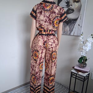 May include: A two-piece outfit comprising a collared, short-sleeved top and wide-leg trousers. The ensemble features a floral pattern in brown, yellow, and burgundy. Black stripes adorn the sleeves, waist, and trouser cuffs. The outfit is displayed on a mannequin.