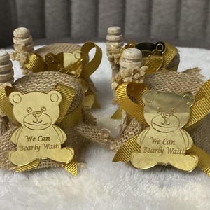 May include: Four small, decorative favors. Each favor features a gold-colored teddy bear charm with the text "We Can Bearly Wait!" attached to a burlap-wrapped item, accented with a gold ribbon and a wooden stopper.