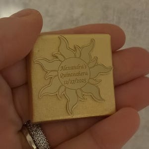 Puede incluir: Un cuadrado dorado con un diseño de sol y la inscripción "Alexandra's Quinceañera 12/27/2025". El diseño está grabado en la superficie. El artículo se sostiene en una mano.