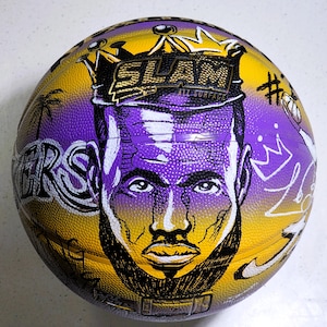 May include: A basketball with a yellow and purple design featuring a portrait of a person with a beard and crown. The ball has the word "SLAM" and "ALL SURFACE" printed on it. The word "LAKERS" is also visible.