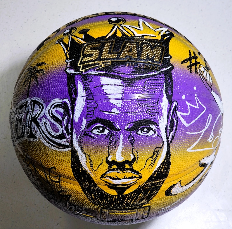 May include: A basketball with a yellow and purple design featuring a portrait of a person with a beard and crown. The ball has the word "SLAM" and "ALL SURFACE" printed on it. The word "LAKERS" is also visible.