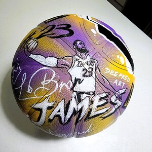 May include: A basketball with a purple, yellow, and black design. The ball features a hand-drawn illustration of a basketball player, the number 23, and the name "JAMES". The words "Dee Fred Art" are also visible.