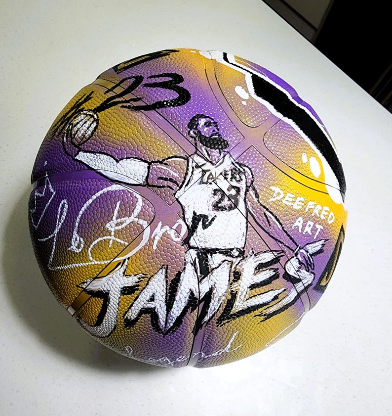 May include: A basketball with a purple, yellow, and black design. The ball features a hand-drawn illustration of a basketball player, the number 23, and the name "JAMES". The words "Dee Fred Art" are also visible.