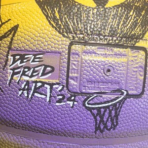 May include: Close-up of a basketball with a textured surface in yellow and purple. The ball has a black illustration and the text "DEE FRED ARTS 24". A small basketball hoop graphic is also visible.