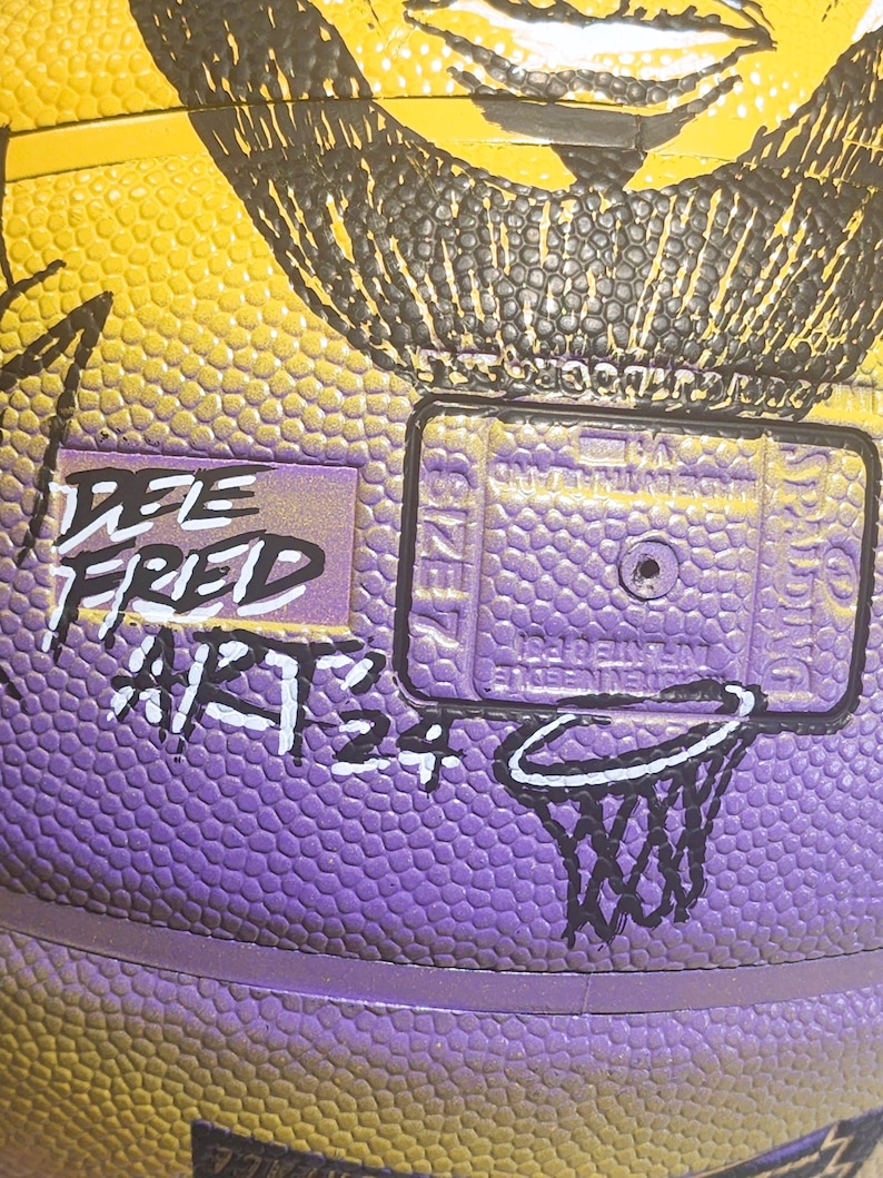 May include: Close-up of a basketball with a textured surface in yellow and purple. The ball has a black illustration and the text "DEE FRED ARTS 24". A small basketball hoop graphic is also visible.
