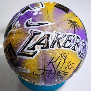 May include: A basketball in the colours yellow, purple, and white, with the word "Lakers" in large letters. The Nike logo, a palm tree, and the words "King James" are also visible on the ball's surface.