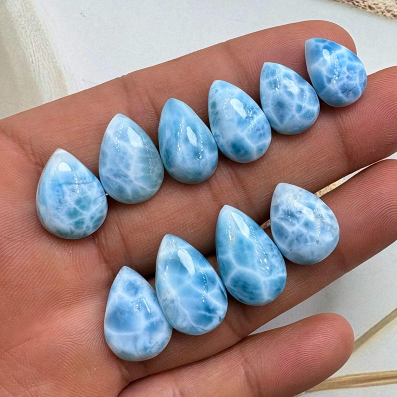 May include: Ten teardrop-shaped larimar gemstones are displayed. The stones are a light blue hue with white marbling, and a polished finish. The gemstones are arranged to highlight their natural beauty and unique patterns. Each stone is approximately 2 cm in length.