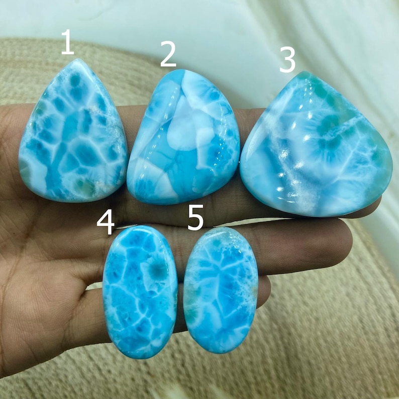 Pu&ograve; includere: Cinque pietre preziose di larimar lucide in varie forme. Le pietre mostrano una gamma di tonalit&agrave; blu con marmorizzazioni bianche, creando un aspetto simile al mare. Le forme includono forme a goccia e ovali, mostrando i modelli naturali.