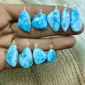 Può includere: Una collezione di nove pietre preziose di larimar lucidate, ognuna con un motivo unico di vortici blu chiaro e bianchi. Le pietre variano nella forma, tra cui goccia, ovale e forme irregolari. Le pietre preziose sono esposte su uno sfondo neutro.