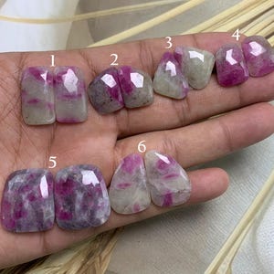 Könnte beinhalten: Sechs Paare von Rubin-Edelstein-Cabochons in verschiedenen Formen. Die Edelsteine haben eine Mischung aus rosa und weißen Farben mit sichtbaren Einschlüssen. Die Cabochons werden auf einer Hand präsentiert und zeigen ihre polierten Oberflächen und einzigartigen Muster.