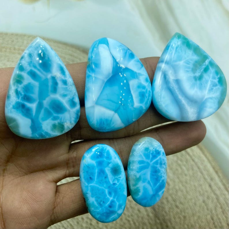 Pu&ograve; includere: Una collezione di pietre preziose di larimar lucide in varie forme, tra cui gocce e ovali. Le pietre mostrano un sorprendente motivo marmorizzato blu e bianco, che ricorda le onde dell'oceano. Le pietre preziose sono tenute in una mano.