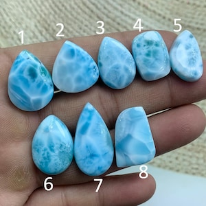 May include: Eight polished larimar gemstones in various shapes, including teardrop and rectangular. The stones display a light blue color with white marbling, resembling ocean waves. The gemstones are held in a hand.
