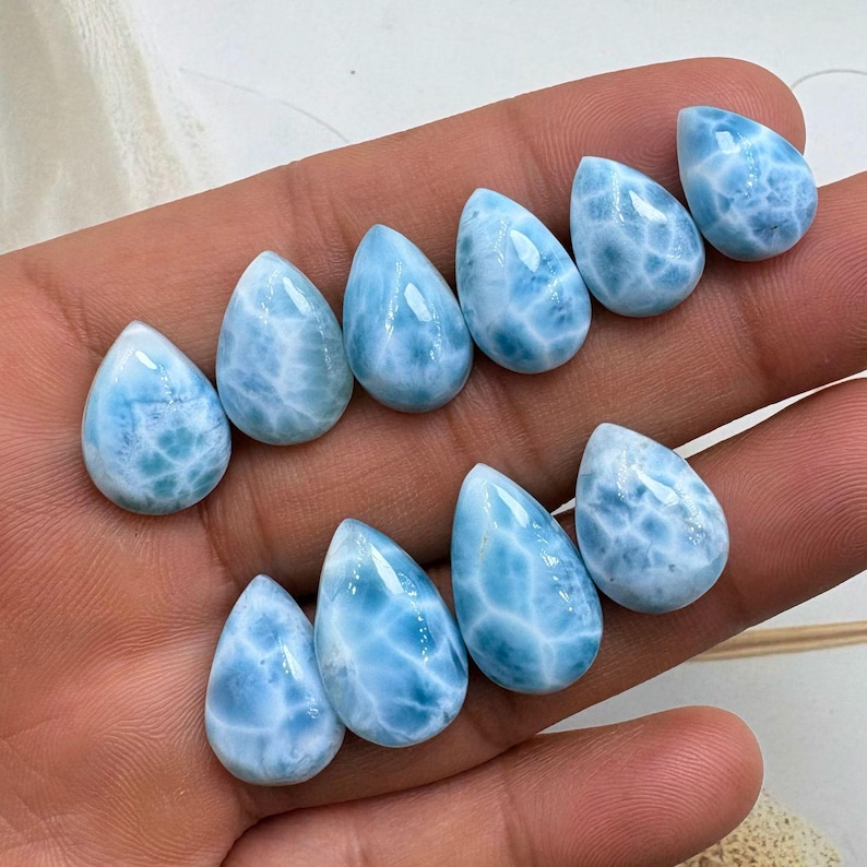 May include: Ten teardrop-shaped larimar gemstones are displayed. The stones are a light blue with white marbling. The polished stones are arranged in two rows, highlighting their unique natural textures and hues. Each stone is approximately 2 cm in length.